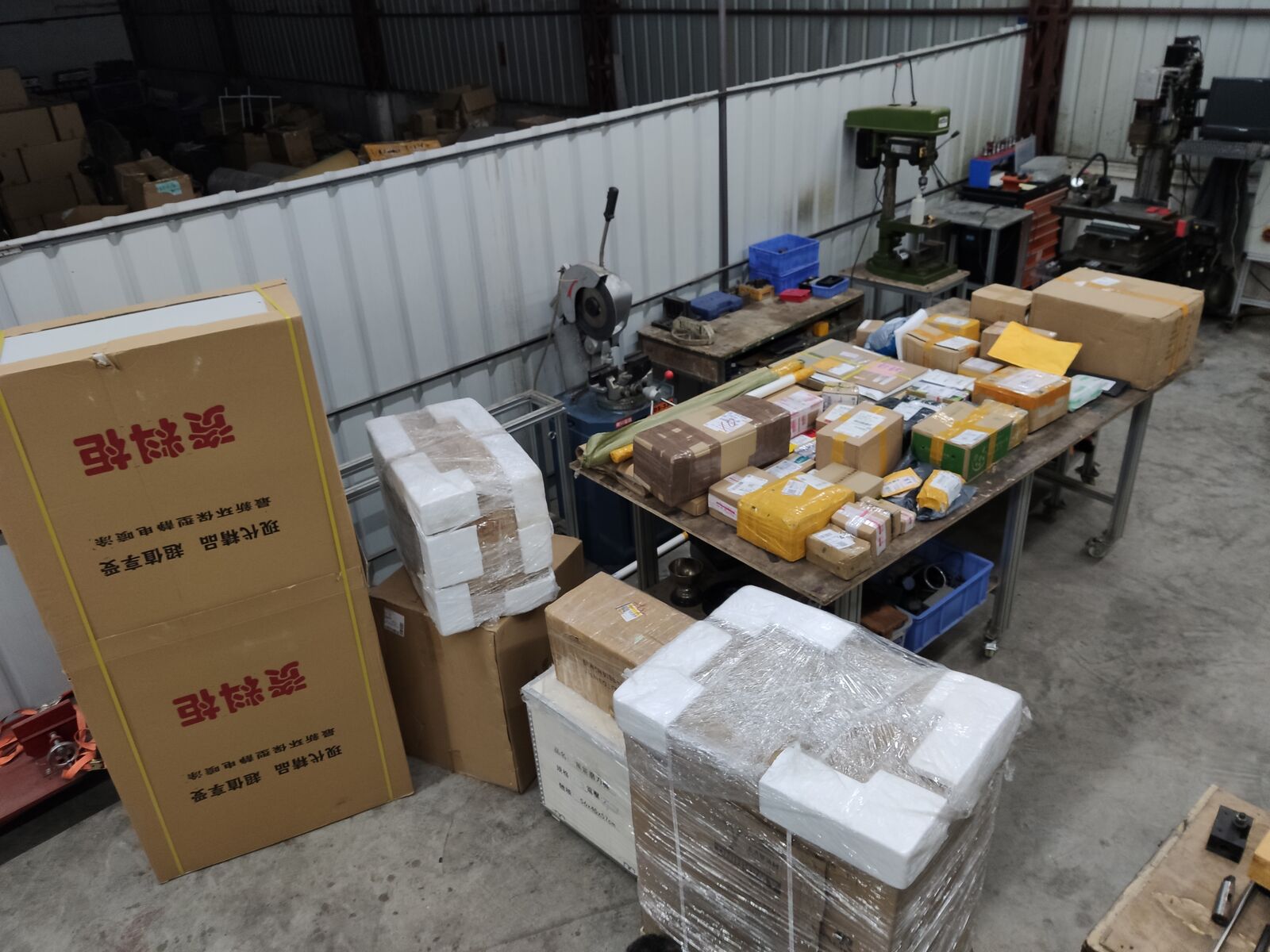 Cardboard parcels and foam-wrapped parts stacked on a workshop table and floor, with a Chinese-labelled box beside them.