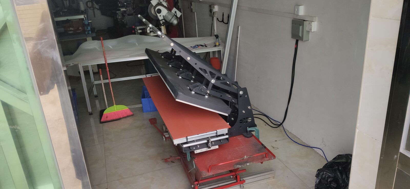 A heat press with its lower bed pulled out in a workshop, with a broom and a window behind.