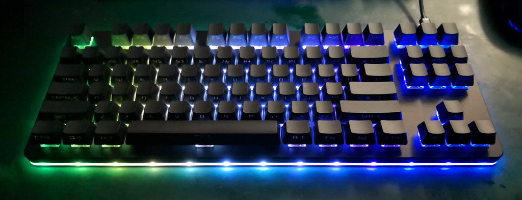 Keyboard with black keycaps and coloured RGB underglow in blue and green along its lower edge.