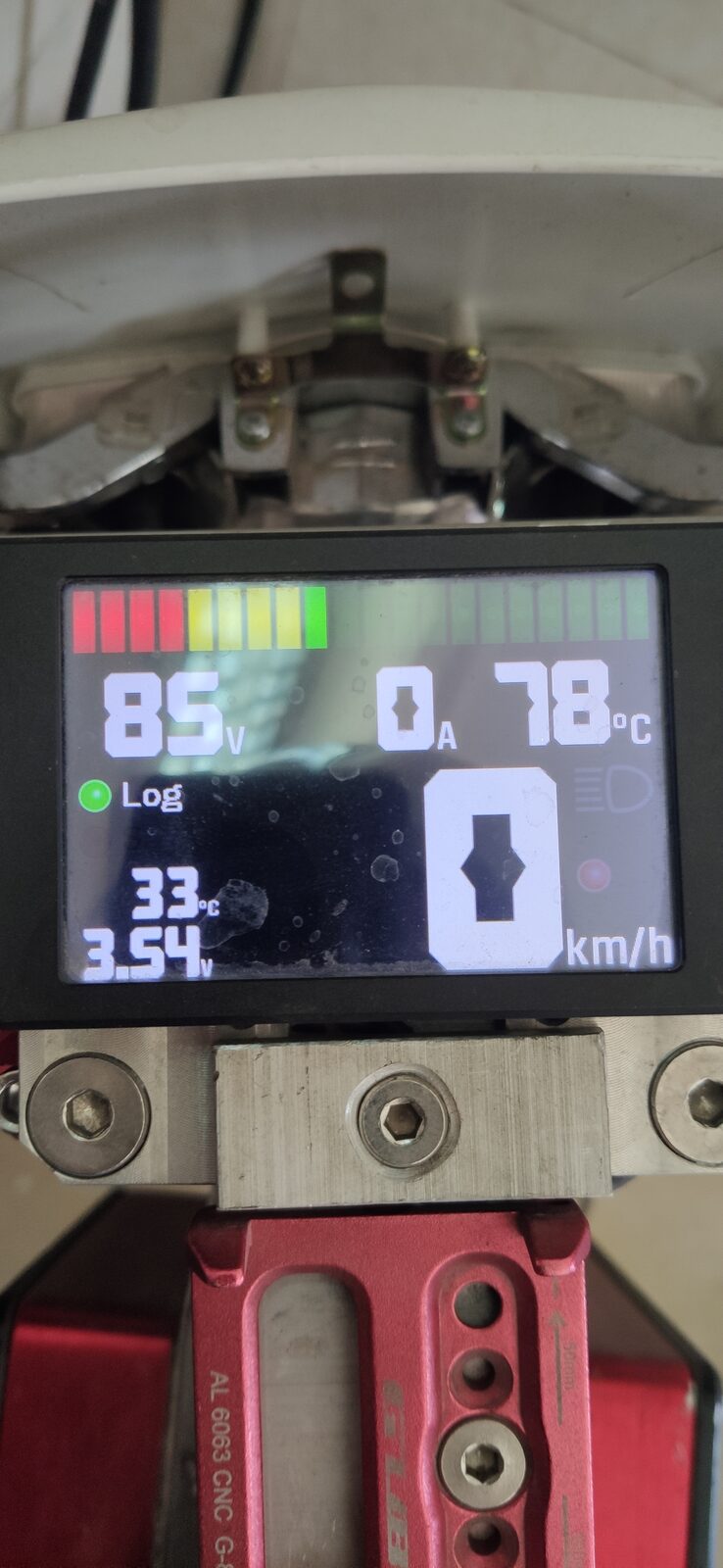 Small colour display showing a battery gauge, numeric readouts and a speed figure, held in a metal bracket with a red part below.
