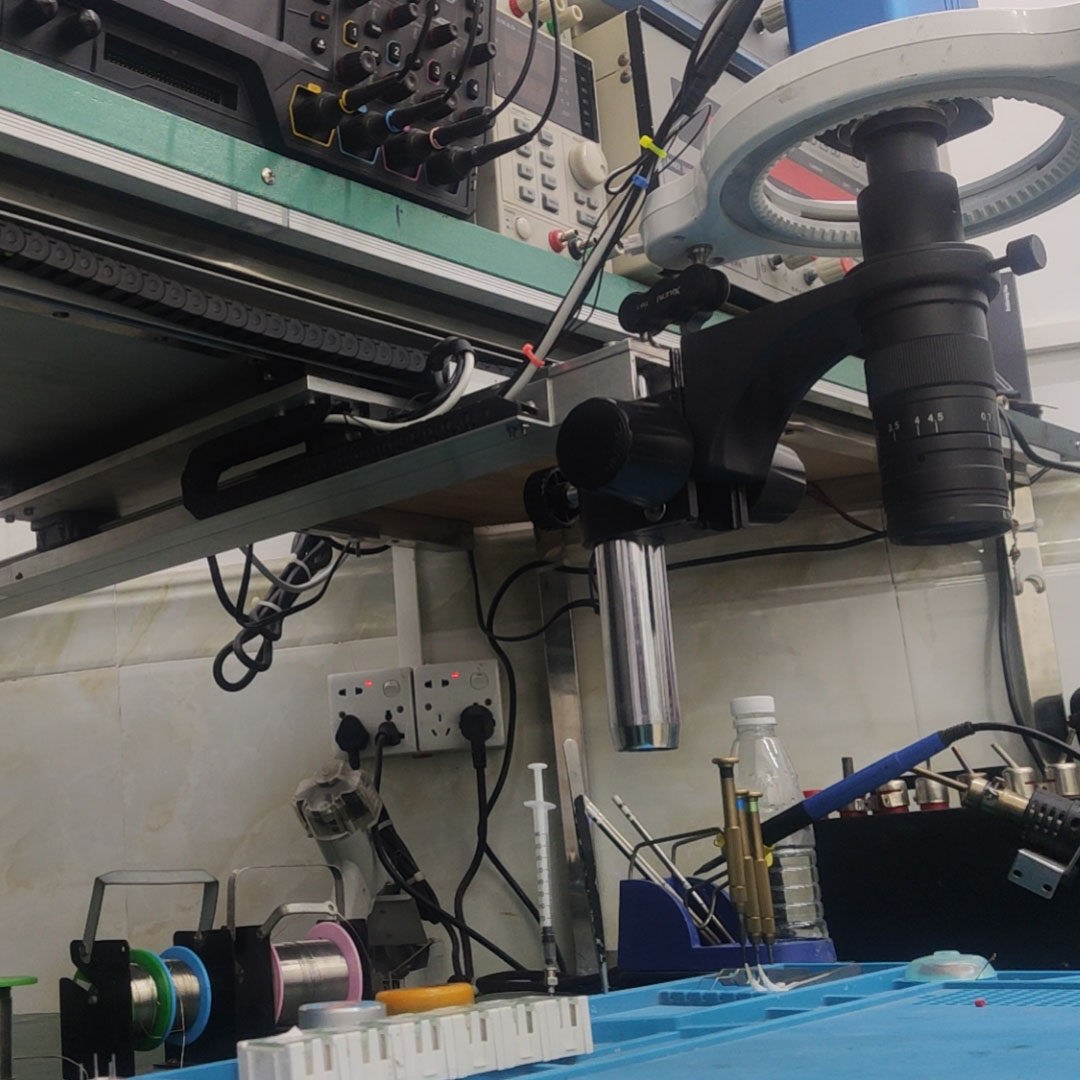 Low-angle view of a ring-lit microscope head on a rail mounted under the bench shelf, with cables and solder spools below.