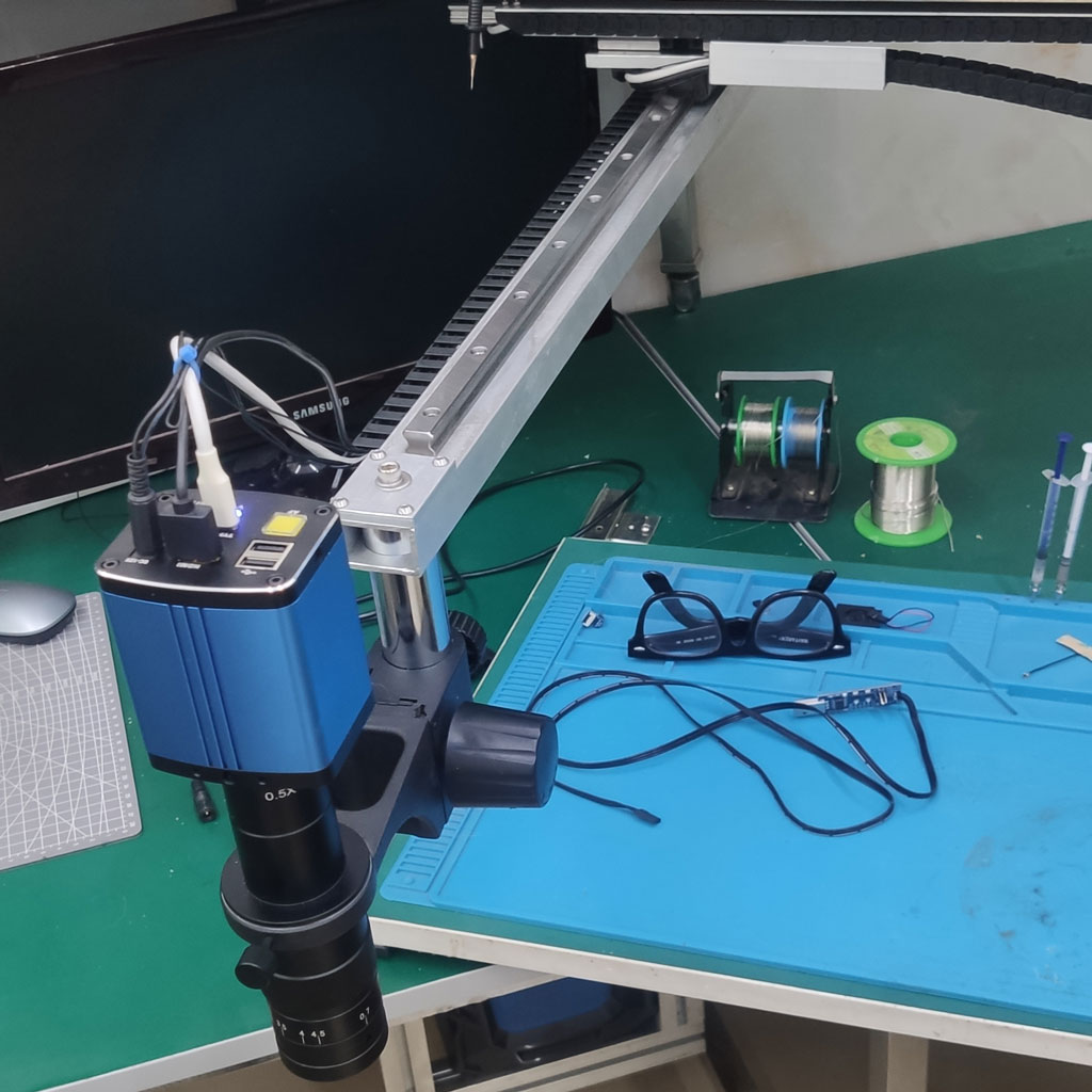 Close view of a blue digital microscope camera hanging from a white sliding rail arm over a blue work mat with glasses, tweezers and cables.